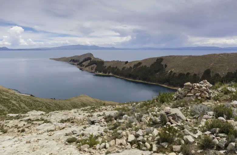 A stone cairn on a dry hillside above a sheltered bay on Lake Titicaca, with distant ranges across the water.