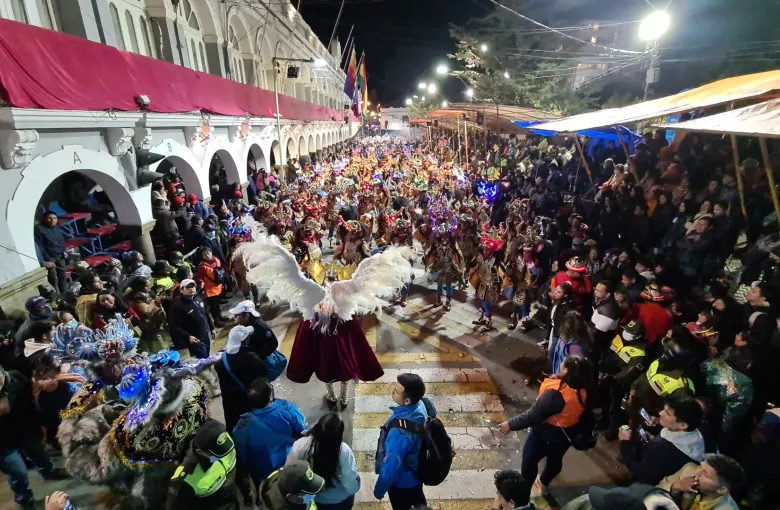 A winged archangel figure leads masked diablada dancers along a street packed with spectators at the Carnival of Oruro.