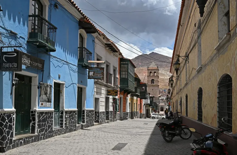 A cobbled Potosi street of blue and ochre colonial houses with wooden balconies, a bell tower and Cerro Rico beyond.