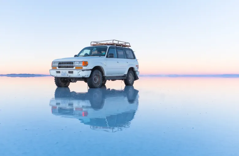 A four wheel drive vehicle stands on the flooded salt flat at dawn, perfectly reflected in the water.