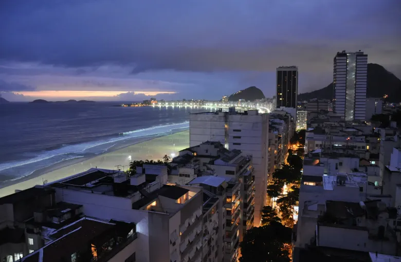 The curve of Copacabana beach at dusk with the lit apartment blocks of the neighbourhood behind it and headlands beyond.