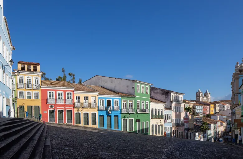 The painted colonial facades of the Largo do Pelourinho in Salvador running down a cobbled slope, with church towers beyond.