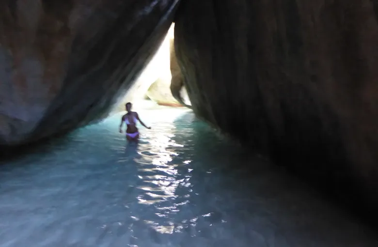 A visitor wading through a narrow sunlit channel between two enormous granite boulders at The Baths.