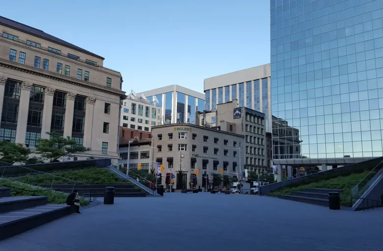 A stone plaza with glass pyramid entrances, the columned Bank of Canada headquarters on one side and downtown Ottawa beyond.