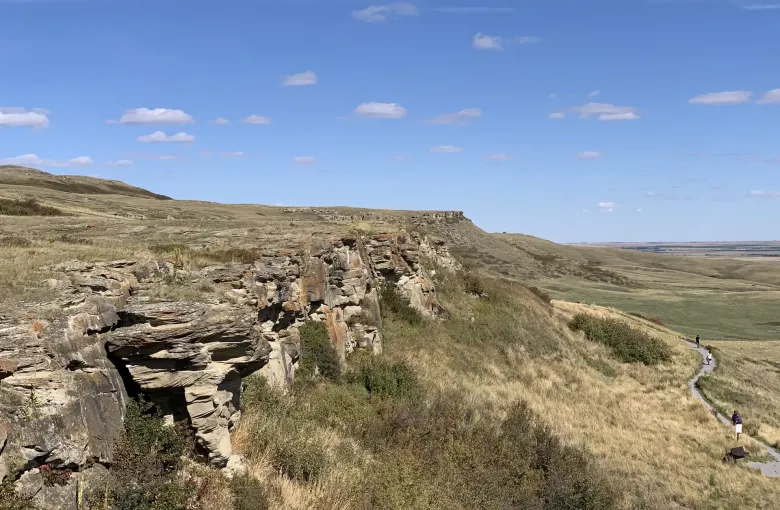 A layered sandstone escarpment above dry grassland, with an interpretive footpath below it and the plains beyond.
