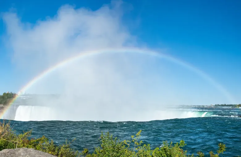 The curved crest of Horseshoe Falls throwing spray high above the gorge, with a full rainbow arcing across the mist.