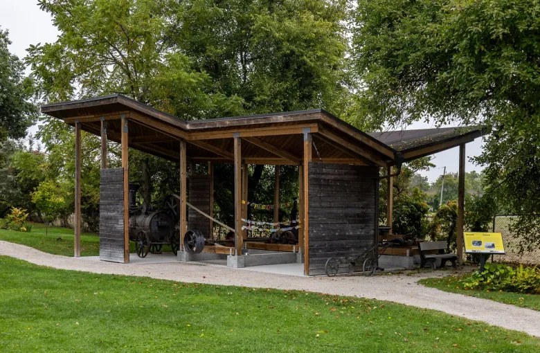 A timber shelter housing a steam engine and sawmill bench, with a bilingual interpretive panel beside the gravel path.