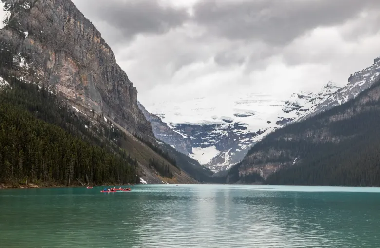 A pale green lake between forested cliffs, a snow and ice field filling the valley head, and red canoes near the shore.