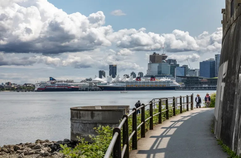 A wide level paved path with a handrail curving along the harbour shore, with walkers on it and city towers beyond.