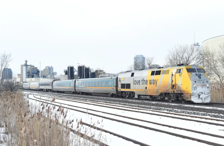 A yellow and silver locomotive hauling passenger cars along snow lined track past low industrial buildings.