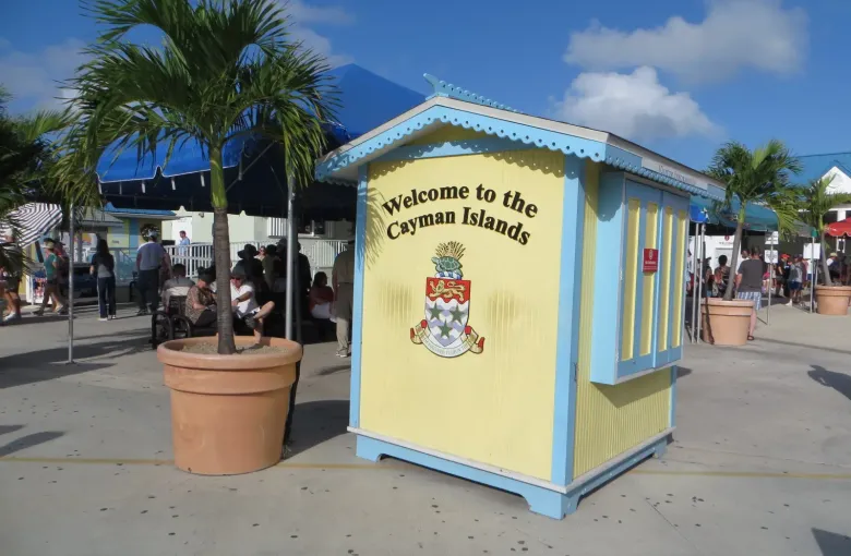A pale blue and yellow kiosk in George Town reading Welcome to the Cayman Islands, with palms and visitors behind.