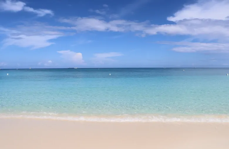 Open sand and shallow turquoise water on Seven Mile Beach under a wide bright sky, looking west to the horizon.