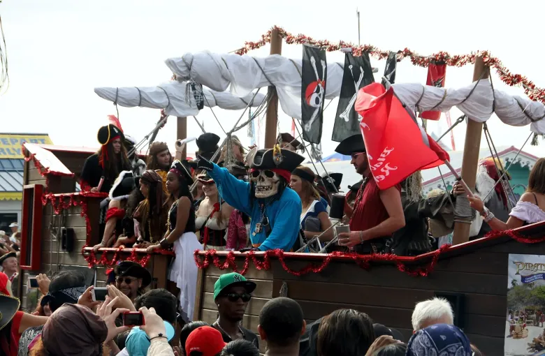 A galleon float with black sails and costumed crew moving through a crowd of spectators during the Pirates Week festival.