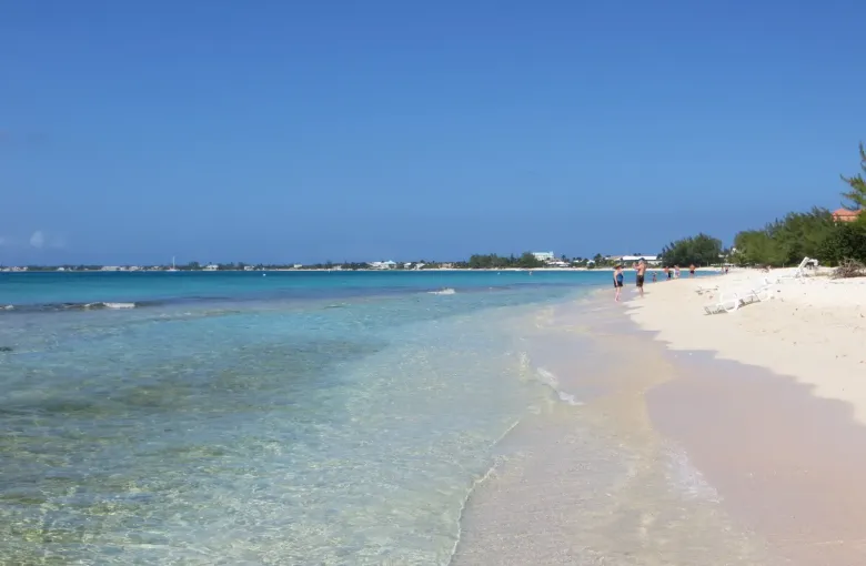 A level stretch of firm pale sand meeting very shallow clear water on Grand Cayman, with people walking far along the shore.