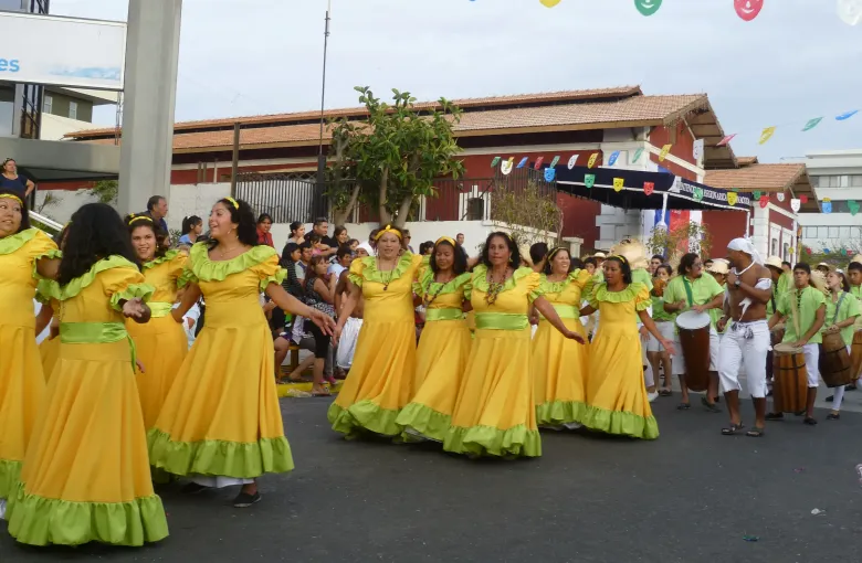 Dancers of the Arica Negro comparsa in yellow and green moving along the street, with drummers carrying tall hand drums behind them.