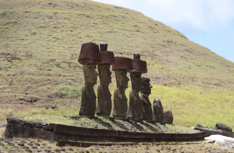 A row of moai standing on their ahu with red stone pukao on their heads, a grass hillside rising behind them.