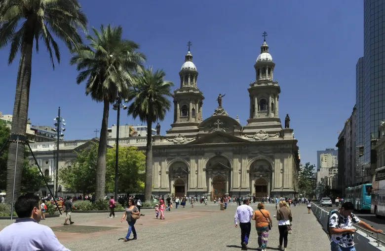 The Metropolitan Cathedral behind palms on the Plaza de Armas in Santiago, with people crossing the square in daylight.