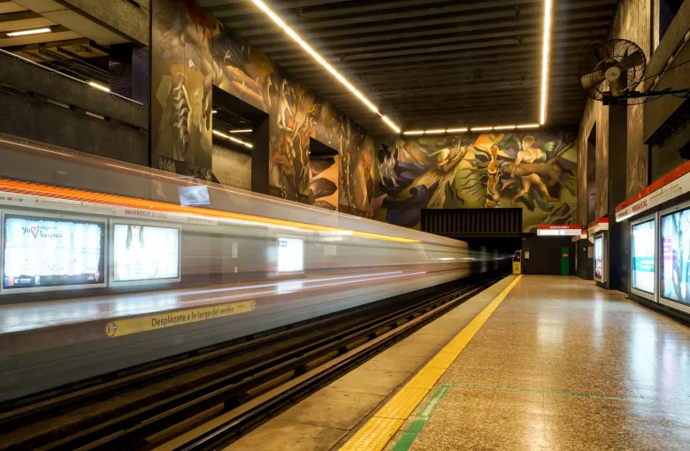 A train blurred with motion along the platform at Universidad de Chile station in Santiago, below the painted mural that lines the wall.