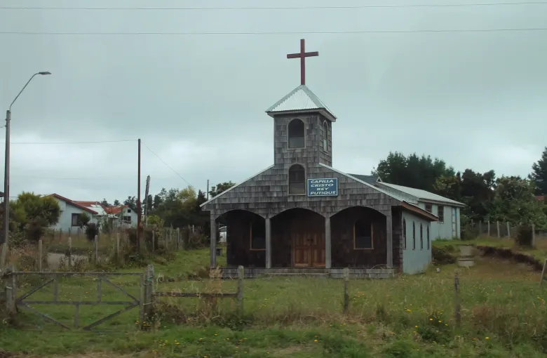 A small chapel clad in wooden shingles with an arcaded porch and a tower, standing in rough grass on the island of Quinchao.