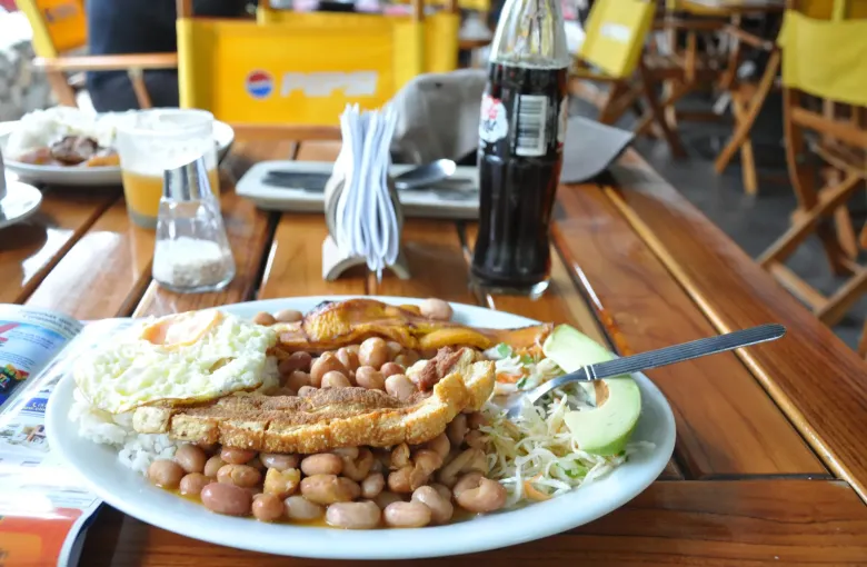 A plate of bandeja paisa on a wooden restaurant table: beans, rice, a fried egg, plantain, chicharron and an arepa.