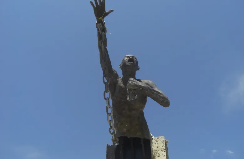 A dark metal statue of a man raising a broken chain above his head, standing against open sky at San Basilio de Palenque.