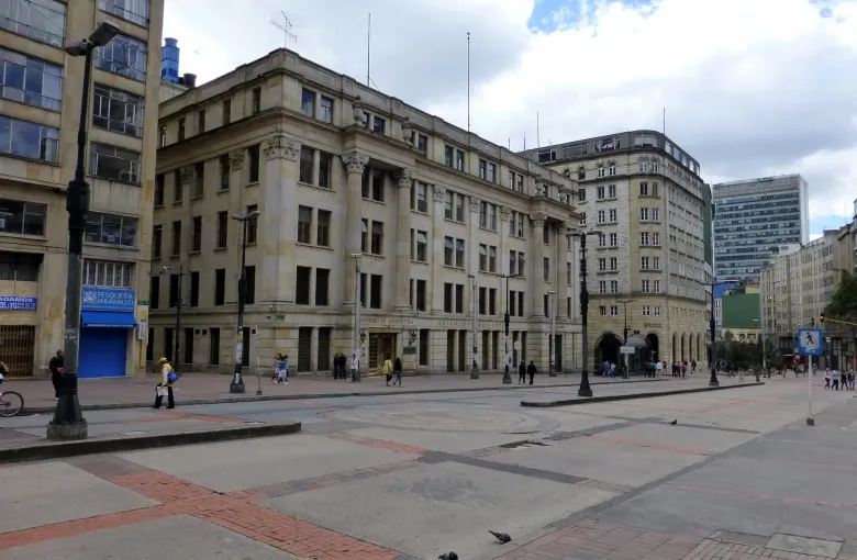 A broad paved square in central Bogota lined with early twentieth century stone buildings, a few people crossing it.