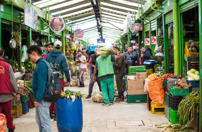 Shoppers and stallholders in a covered market hall in Bogota, stacked crates of vegetables and fruit along both aisles.