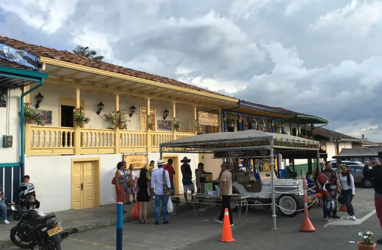 A street in Salento: a long single-storey building with a painted timber balcony and clay-tile roof, jeeps and people outside it, mountains behind.