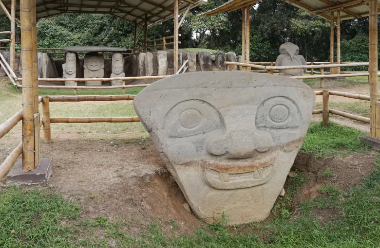 A large carved stone figure with a broad square face at the San Agustin archaeological park, standing under a protective shelter.