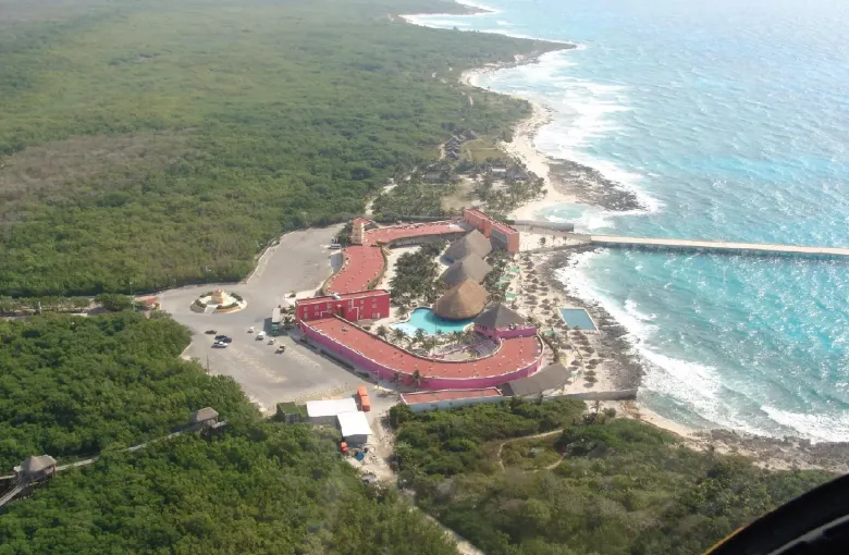An aerial view of the cruise terminal buildings on a low green shore, with surf breaking along a reef just offshore.