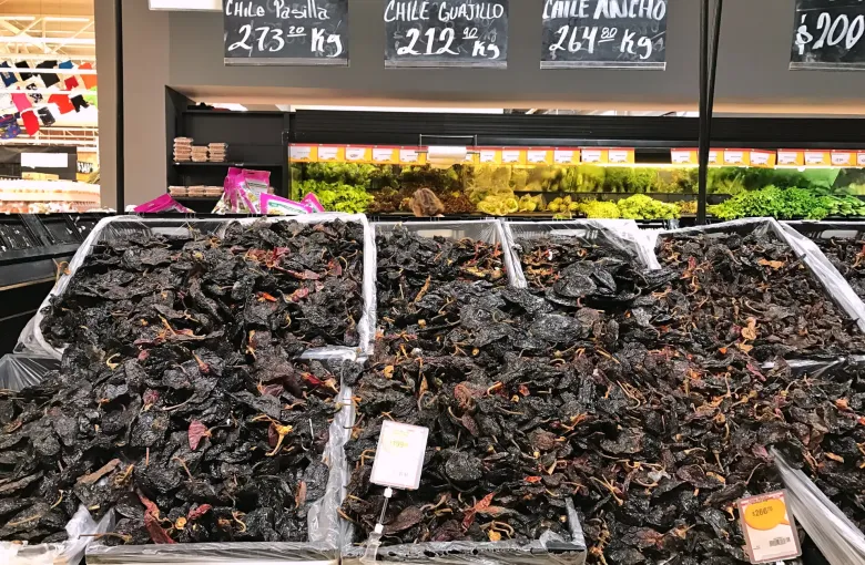 Trays of dried chiles in a market, each with a chalkboard giving its name and a price per kilo in pesos.