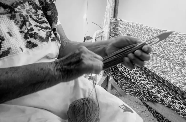 Hands at work on a backstrap loom in black and white, drawing a shuttle through the warp beside a ball of yarn.