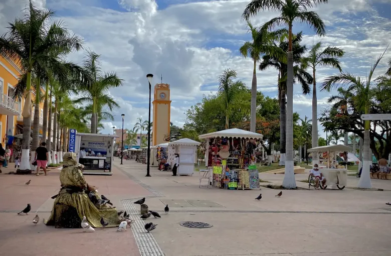 A palm-lined town square with market stalls under white canopies, a bronze figure on a plinth and a clock tower.