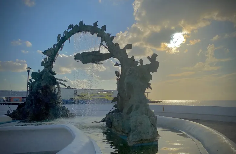 A dark bronze sculpture arching over a circular fountain on the seafront at dusk, with open water beyond.