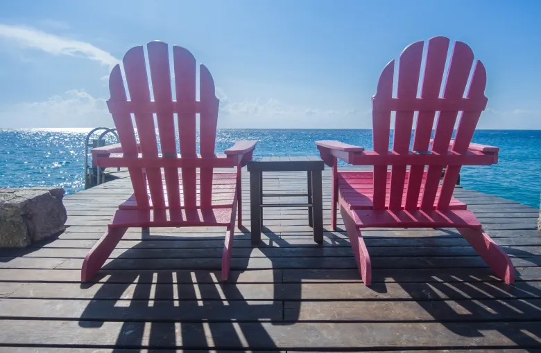 Two pink wooden deck chairs and a small table on a timber pier, facing bright turquoise water under a blue sky.
