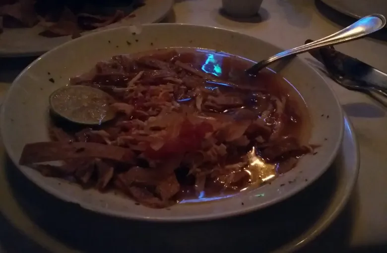 A shallow bowl of sopa de lima: shredded chicken and tortilla strips in clear broth with a slice of lime.