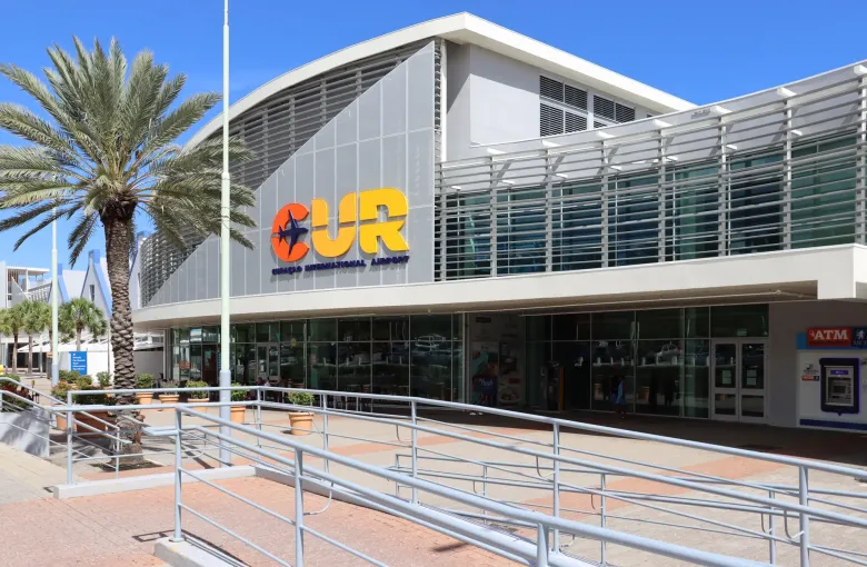 The terminal frontage of Curaçao International Airport, with its CUR sign, a ramped walkway and palms against a clear sky.