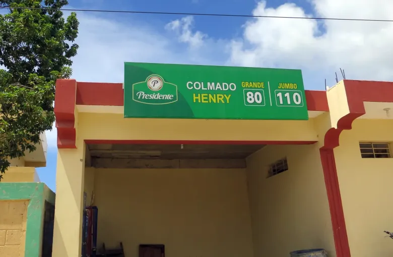 A Dominican colmado with a painted sign above the doorway listing two prices, under a blue sky.