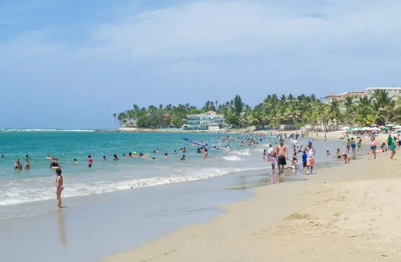 People swim and walk along a curving north coast beach backed by palms and low buildings.