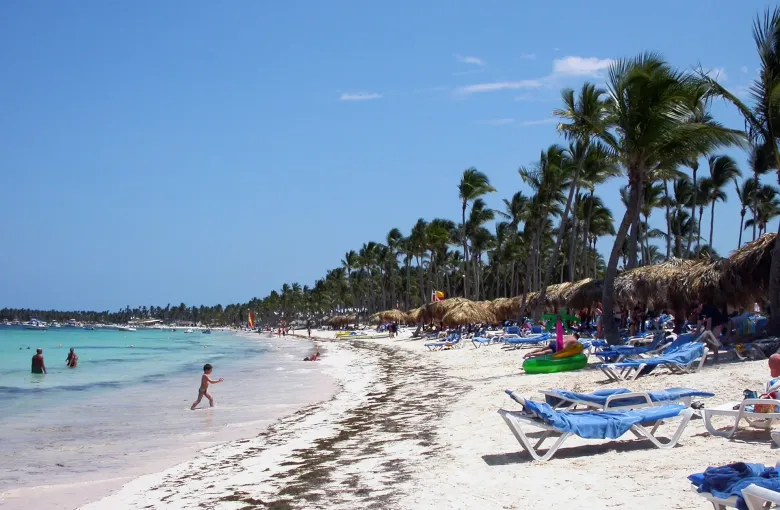Loungers and thatched shades line a palm-backed beach at Punta Cana, with swimmers in the shallows.