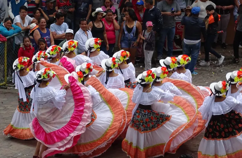 Dancers in wide ruffled skirts and flower-trimmed hats turning together in a street carnival, seen from above.
