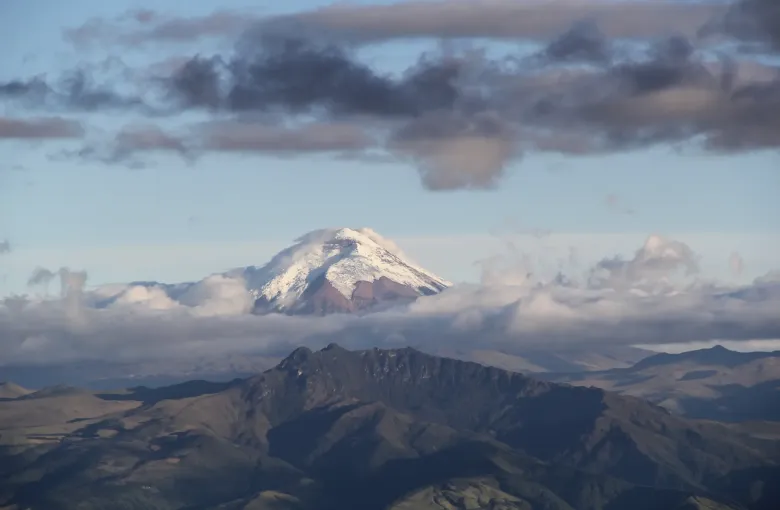 The snow-capped cone of Cotopaxi rising above a band of cloud, with dark Andean ridges in the foreground.