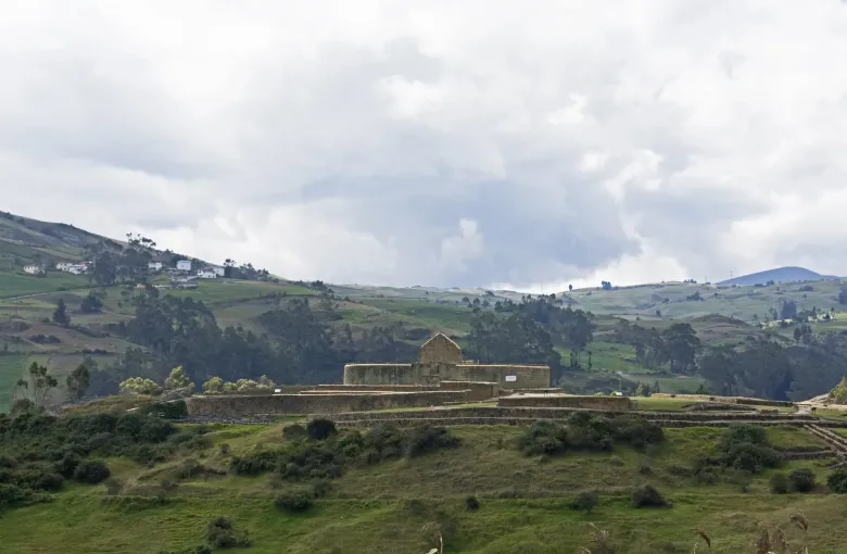 The stone terraces and elliptical temple of Ingapirca standing on green highland pasture under heavy cloud.