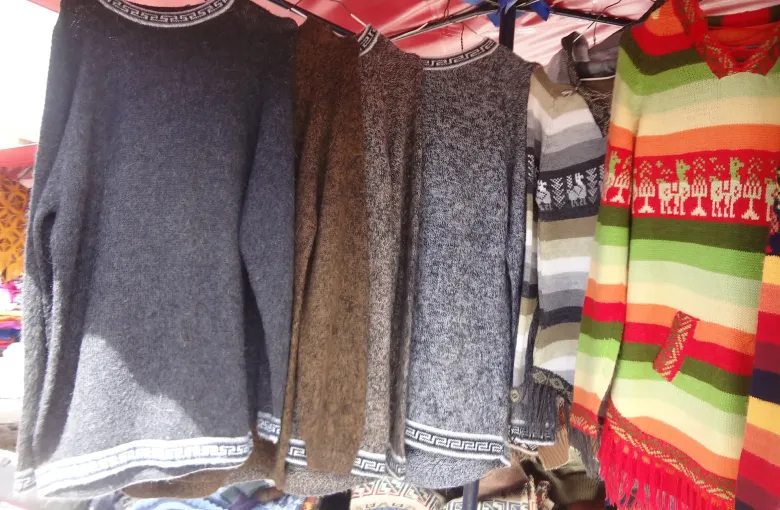 Knitted sweaters and a striped poncho hanging from a market stall frame under a red canopy at Otavalo.