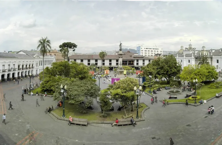 A wide view across Plaza Grande in Quito, showing flat paved walkways between the gardens and the surrounding colonial facades.
