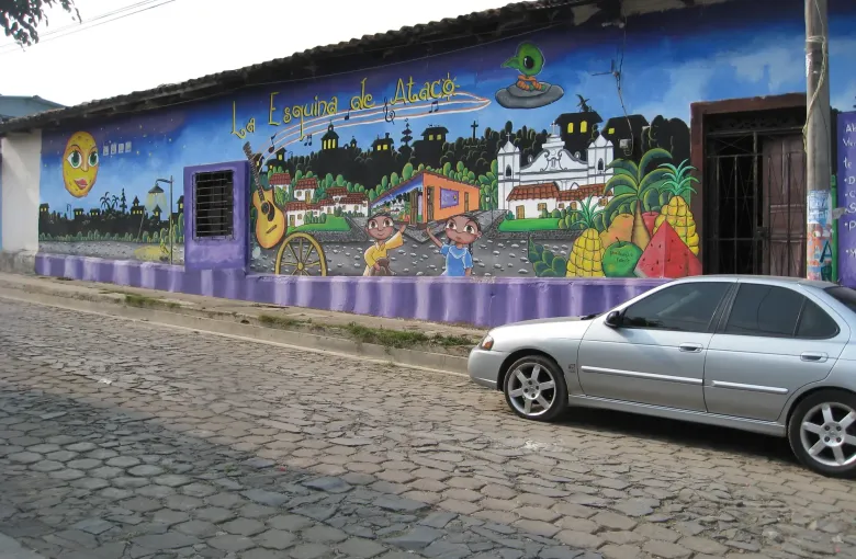 A blue wall painted with a village scene of houses, children and fruit, above a cobbled street with a parked car.