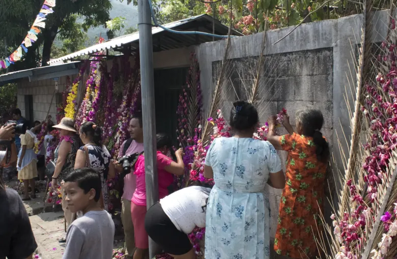 Women and children working along a wall hung with palm fronds, threading bright paper flowers into the leaves.