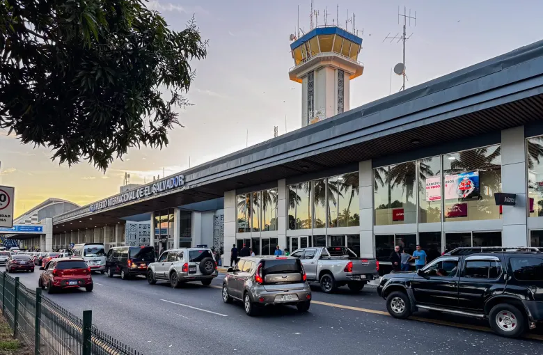 A low airport terminal with a tiled control tower above it and cars queueing along the departures road at dusk.