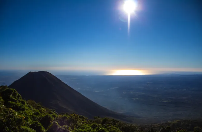 The dark cone of Izalco seen from above at sunset, with the sun low over a hazy plain and the sea beyond.