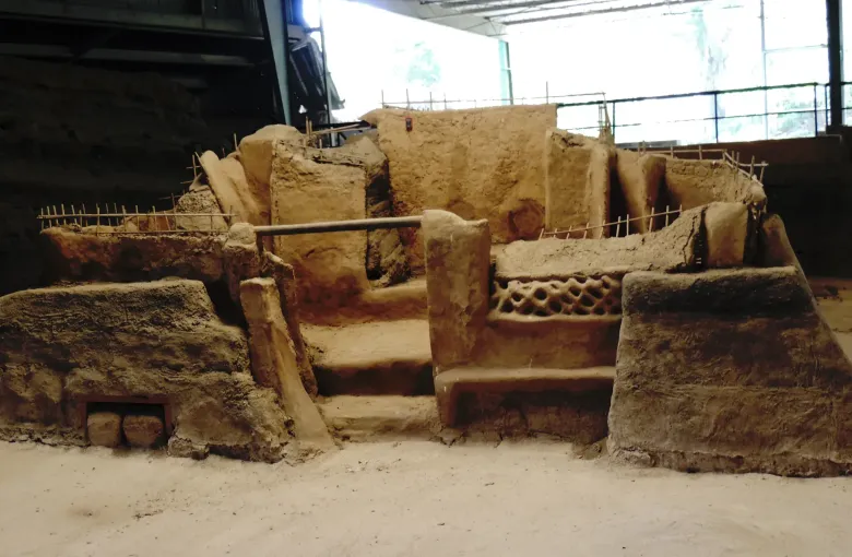 Earthen walls and doorways of a buried village standing in an excavation pit beneath a protective roof.